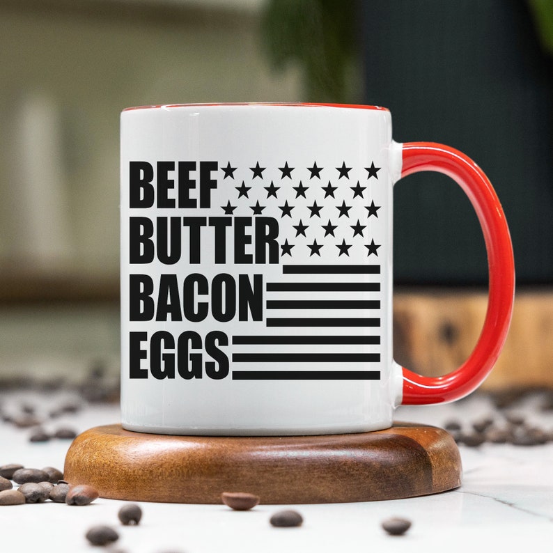 K&ouml;nnte beinhalten: Eine wei&szlig;e Keramiktasse mit rotem Rand und Henkel. Die Tasse hat ein schwarz-wei&szlig;es amerikanisches Flaggenmuster mit dem Aufdruck "BEEF BUTTER BACON EGGS".