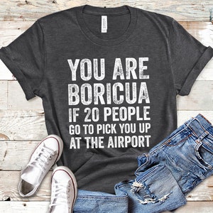 Puede incluir: Camiseta gris oscuro con el texto blanco "YOU ARE BORICUA IF 20 PEOPLE GO TO PICK YOU UP AT THE AIRPORT". La camiseta se combina con vaqueros azules y zapatillas blancas.