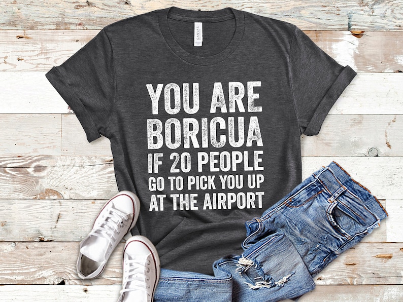 Puede incluir: Camiseta gris oscuro con el texto blanco "YOU ARE BORICUA IF 20 PEOPLE GO TO PICK YOU UP AT THE AIRPORT". La camiseta se combina con vaqueros azules y zapatillas blancas.