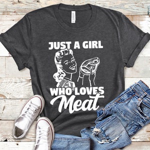 May include: A dark gray t-shirt with a white graphic of a woman holding a steak and the text "Just a girl who loves meat".