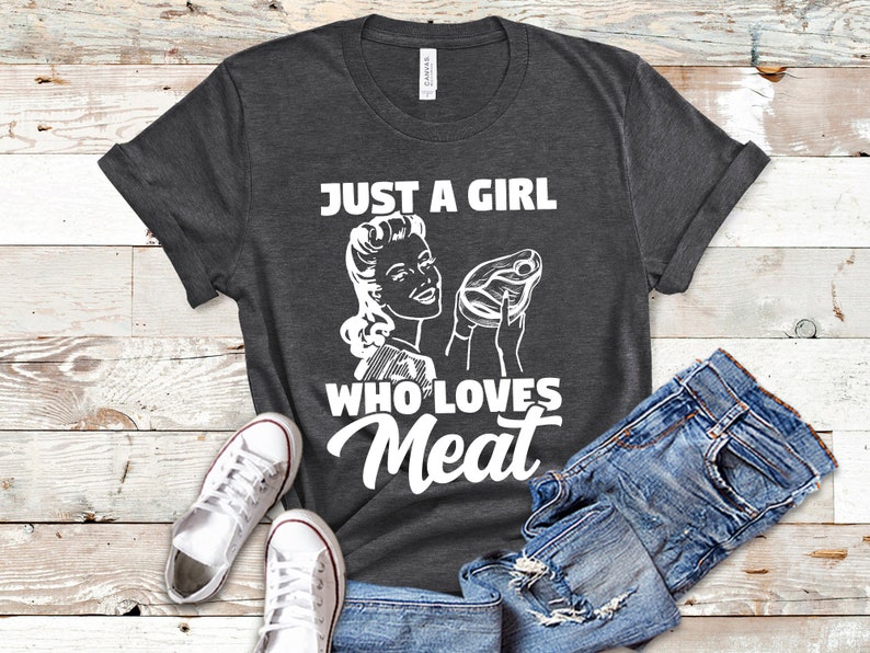 May include: A dark gray t-shirt with a white graphic of a woman holding a steak and the text "Just a girl who loves meat".