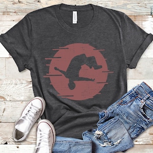 May include: A dark heather grey t-shirt with a silhouette of a person doing a backflip in a red circle with white streaks.