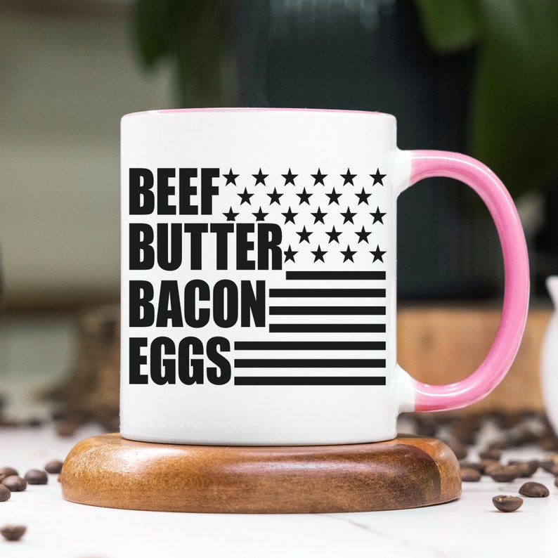 K&ouml;nnte beinhalten: Wei&szlig;e Keramik-Kaffeetasse mit rosafarbenem Rand und Henkel. Die Tasse zeigt ein schwarz-wei&szlig;es amerikanisches Flaggenmuster mit dem Aufdruck "BEEF BUTTER BACON EGGS".