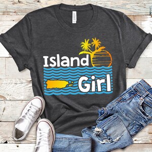 May include: A dark gray t-shirt with the text "Island Girl" in white and blue. The design features a yellow silhouette of Puerto Rico, palm trees, and a sunset.
