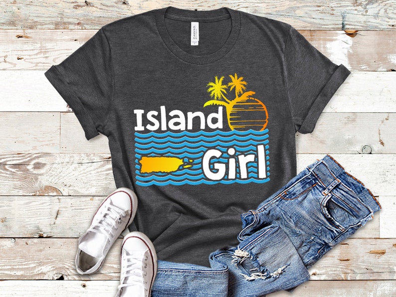 May include: A dark gray t-shirt with the text "Island Girl" in white and blue. The design features a yellow silhouette of Puerto Rico, palm trees, and a sunset.