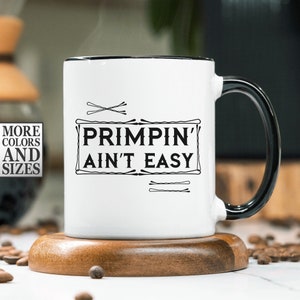May include: White ceramic mug with a black rim and handle. The mug features the text "PRIMPIN' AIN'T EASY" in black, framed by decorative elements. The mug sits on a wooden coaster, with coffee beans scattered around.