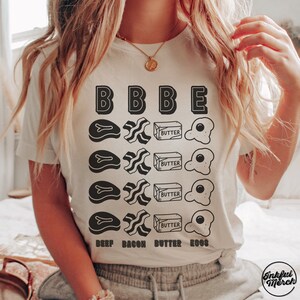 May include: White t-shirt with a graphic design of a breakfast theme. The graphic includes the words "BEEF BACON BUTTER EGGS" and a series of illustrations of beef, bacon, butter, and eggs.