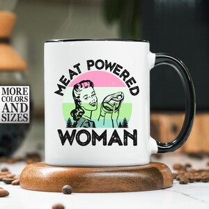 Può includere: Tazza in ceramica bianca con manico e bordo neri. La tazza presenta la scritta "MEAT POWERED WOMAN" con un'illustrazione retrò di una donna che tiene una bistecca. La tazza è su un sottobicchiere di legno.