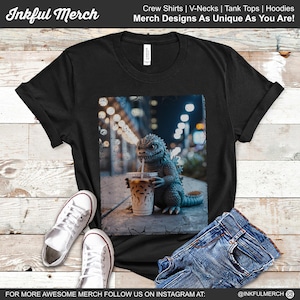 May include: Black t-shirt featuring a Godzilla figure drinking iced coffee with a straw. The graphic is set against a blurred city background. The shirt is paired with white sneakers and blue jeans. Text on the shirt says "Merch Designs As Unique As You Are!"