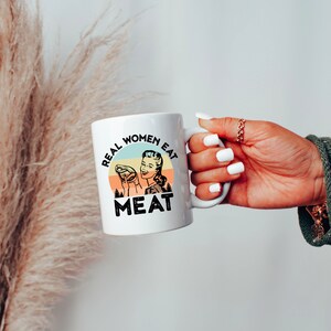 May include: White ceramic mug with a retro-style illustration of a woman holding a piece of meat. The text on the mug reads "Real Women Eat Meat".
