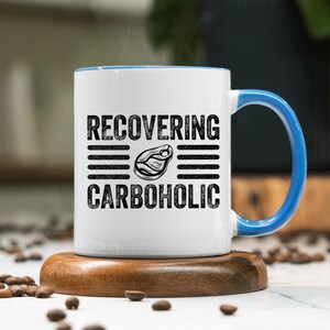 May include: White ceramic mug with a blue rim and handle. The mug has black text that reads "RECOVERING CARBOHOLIC" with a black and white illustration of a steak in between the words.
