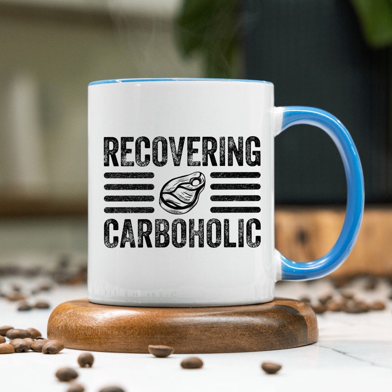 May include: White ceramic mug with a blue rim and handle. The mug has black text that reads "RECOVERING CARBOHOLIC" with a black and white illustration of a steak in between the words.