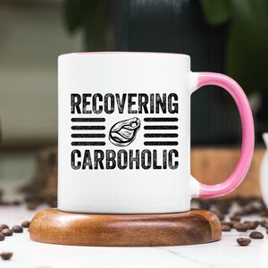 May include: White ceramic mug with a pink rim and handle. The mug has black text that reads "RECOVERING CARBOHOLIC" with a black and white illustration of a steak in between the words.