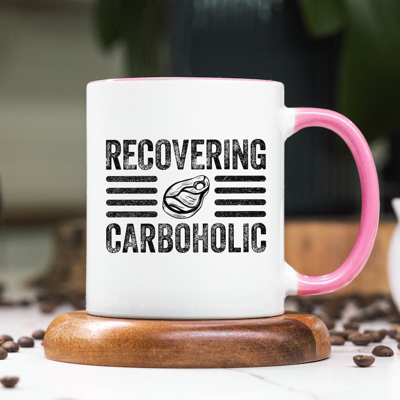 May include: White ceramic mug with a pink rim and handle. The mug has black text that reads "RECOVERING CARBOHOLIC" with a black and white illustration of a steak in between the words.