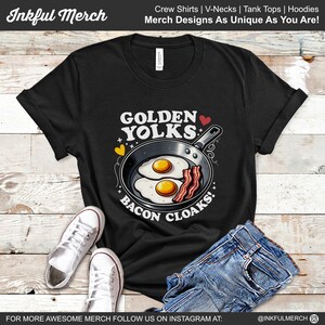 May include: Black t-shirt with a graphic of a frying pan with two sunny-side-up eggs and bacon. The text "Golden Yolks" and "Bacon Cloaks!" is printed on the shirt.