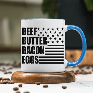 K&ouml;nnte beinhalten: Eine wei&szlig;e Keramik-Kaffeetasse mit blauem Rand und Henkel. Die Tasse hat ein schwarz-wei&szlig;es amerikanisches Flaggenmuster mit dem Aufdruck "BEEF BUTTER BACON EGGS".