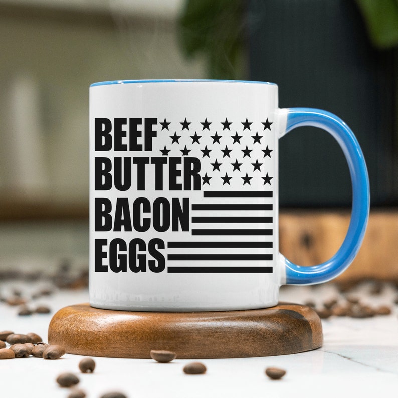 K&ouml;nnte beinhalten: Eine wei&szlig;e Keramik-Kaffeetasse mit blauem Rand und Henkel. Die Tasse hat ein schwarz-wei&szlig;es amerikanisches Flaggenmuster mit dem Aufdruck "BEEF BUTTER BACON EGGS".