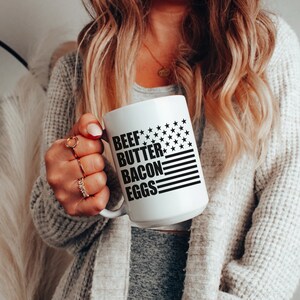 K&ouml;nnte beinhalten: Eine wei&szlig;e Keramiktasse mit einem schwarz-wei&szlig;en amerikanischen Flaggenmuster. Der Text "BEEF BUTTER BACON EGGS" ist auf die Tasse gedruckt.