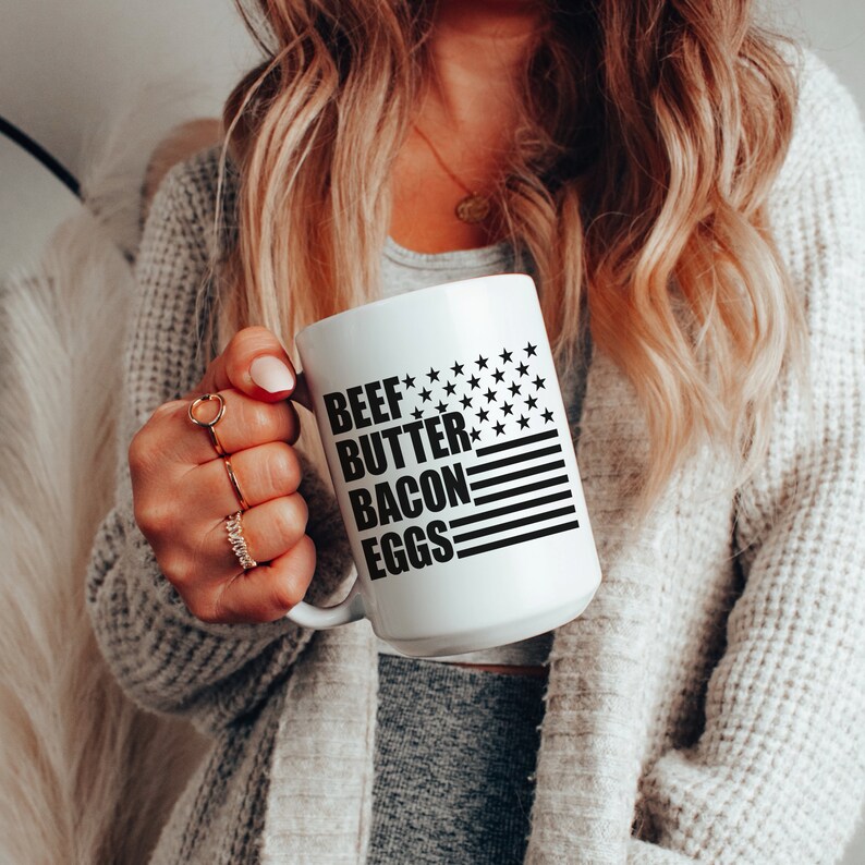 K&ouml;nnte beinhalten: Eine wei&szlig;e Keramiktasse mit einem schwarz-wei&szlig;en amerikanischen Flaggenmuster. Der Text "BEEF BUTTER BACON EGGS" ist auf die Tasse gedruckt.