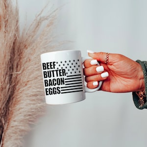 K&ouml;nnte beinhalten: Eine wei&szlig;e Keramik-Kaffeetasse mit einem schwarz-wei&szlig;en amerikanischen Flaggenmuster und dem Text "BEEF BUTTER BACON EGGS" auf der Vorderseite.