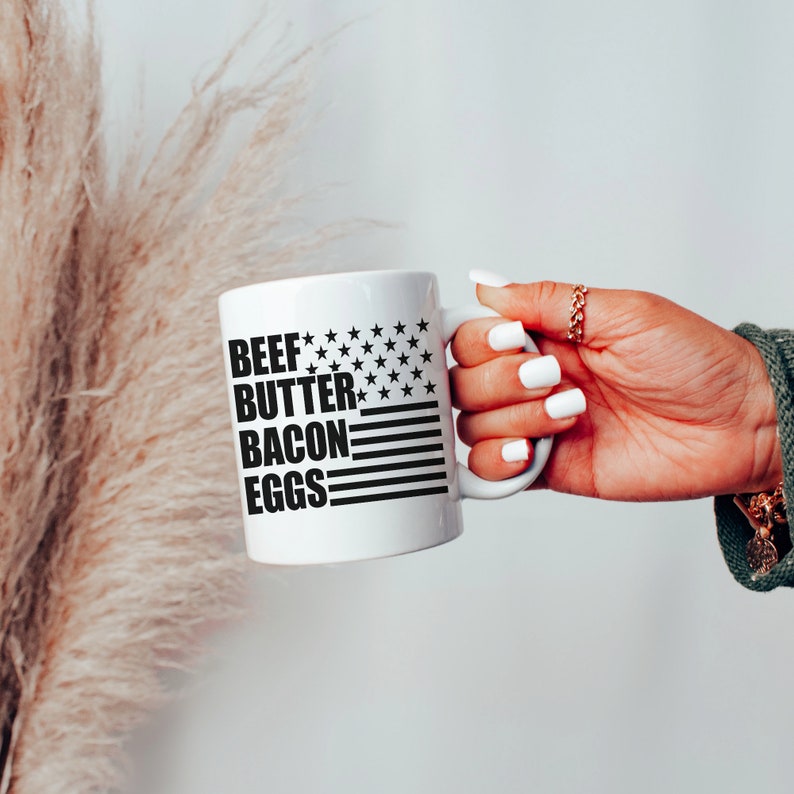 K&ouml;nnte beinhalten: Eine wei&szlig;e Keramik-Kaffeetasse mit einem schwarz-wei&szlig;en amerikanischen Flaggenmuster und dem Text "BEEF BUTTER BACON EGGS" auf der Vorderseite.