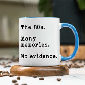May include: White ceramic mug with a blue rim and handle. The mug has black text that reads "The 80s. Many memories. No evidence."