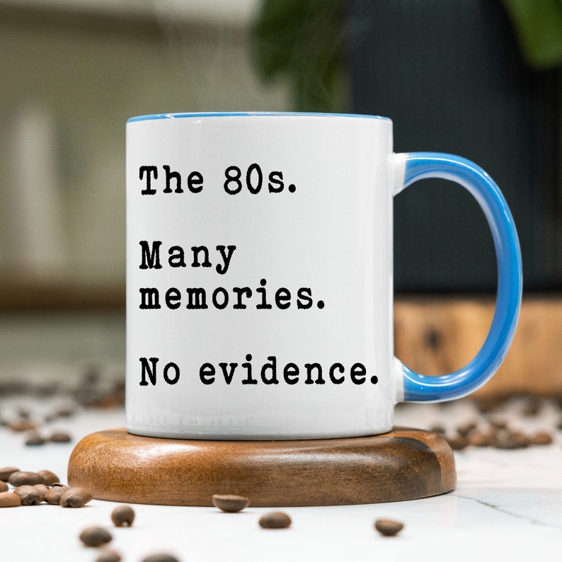 May include: White ceramic mug with a blue rim and handle. The mug has black text that reads "The 80s. Many memories. No evidence."