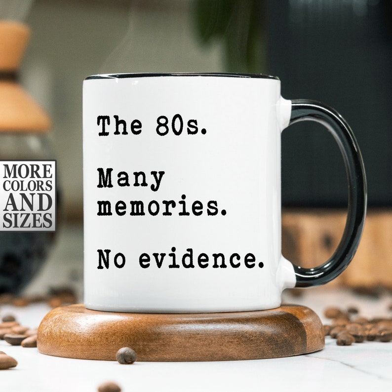 May include: White ceramic mug with a black handle and black rim. The mug has black text that reads "The 80s. Many memories. No evidence."
