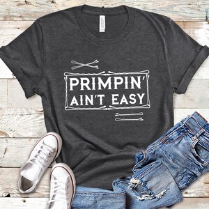 May include: A dark gray t-shirt with the text "Primpin' Ain't Easy" in white lettering. The text is framed by a rectangle with hair clips at the top and bottom.