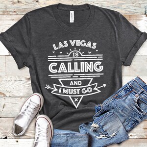 Pu&ograve; includere: Una maglietta grigia scura con testo bianco che recita "Las Vegas is Calling and I Must Go". Il testo &egrave; stilizzato con un sole, frecce e cuori.