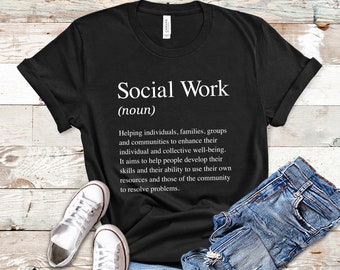 May include: Black t-shirt with white text that reads "Social Work (noun)" and a definition of the term. The shirt is laid flat on a wooden surface with a pair of white sneakers and blue jeans.