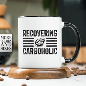 May include: White ceramic mug with a black handle. The mug has black text that reads "RECOVERING CARBOHOLIC" with a black and white illustration of a steak in between the words.