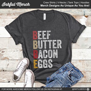 May include: A dark gray t-shirt with the words "BEEF BUTTER BACON EGGS" printed in red, yellow, and white on the front. The shirt is laid flat on a wooden surface with a pair of white sneakers and blue jeans.