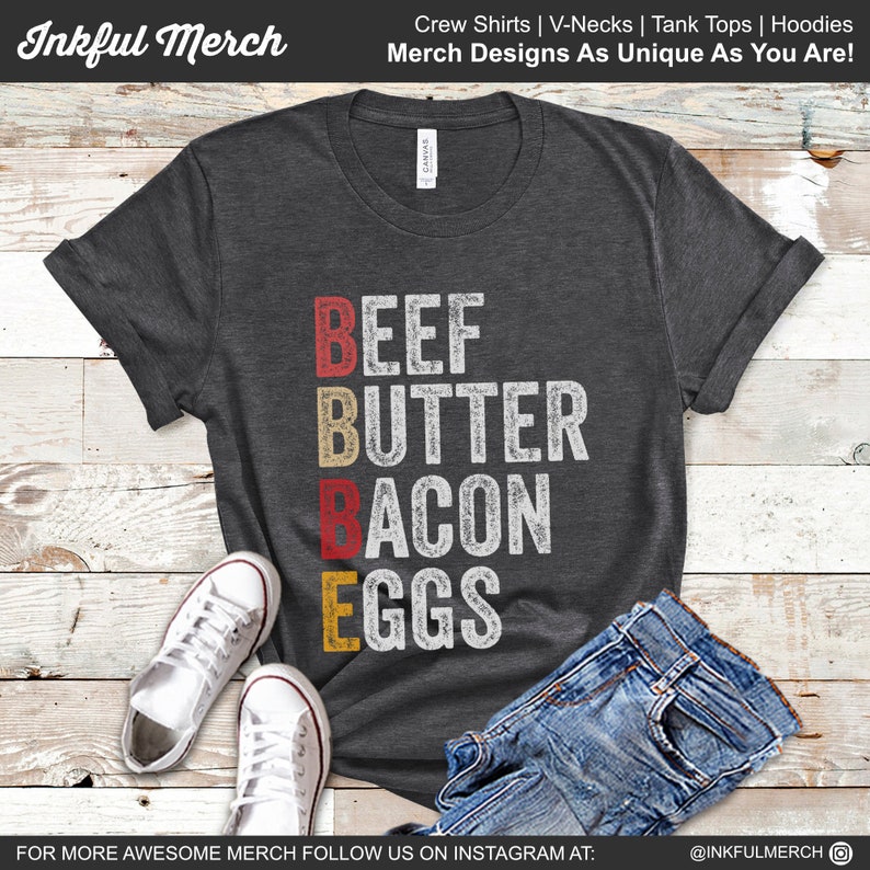 May include: A dark gray t-shirt with the words "BEEF BUTTER BACON EGGS" printed in red, yellow, and white on the front. The shirt is laid flat on a wooden surface with a pair of white sneakers and blue jeans.