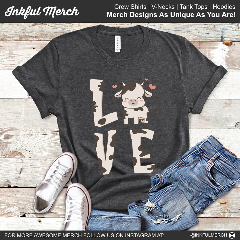 Puede incluir: Una camiseta gris oscuro con un gr&aacute;fico de vaca blanca y la palabra "LOVE" escrita en letras blancas con manchas de vaca negras. La vaca lleva un coraz&oacute;n rojo en la cabeza y hay dos corazones rojos sobre la vaca. La camiseta est&aacute; colocada sobre una superficie de madera con un par de zapatillas blancas y un par de pantalones vaqueros azules.