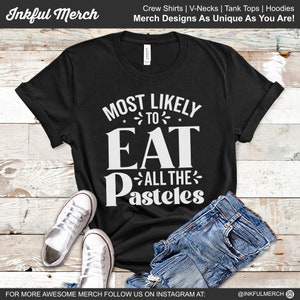 May include: Black t-shirt with white text that reads "Most Likely to Eat All the Pasteles". The shirt is laid flat on a wooden surface with a pair of white sneakers and blue jeans.