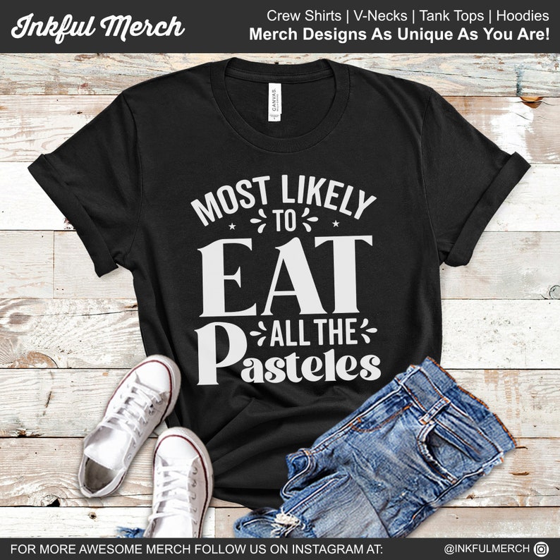 May include: Black t-shirt with white text that reads "Most Likely to Eat All the Pasteles". The shirt is laid flat on a wooden surface with a pair of white sneakers and blue jeans.