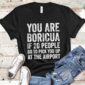 Puede incluir: Camiseta negra con texto blanco: "YOU ARE BORICUA IF 20 PEOPLE GO TO PICK YOU UP AT THE AIRPORT." La camiseta se muestra con vaqueros azules y zapatillas blancas.