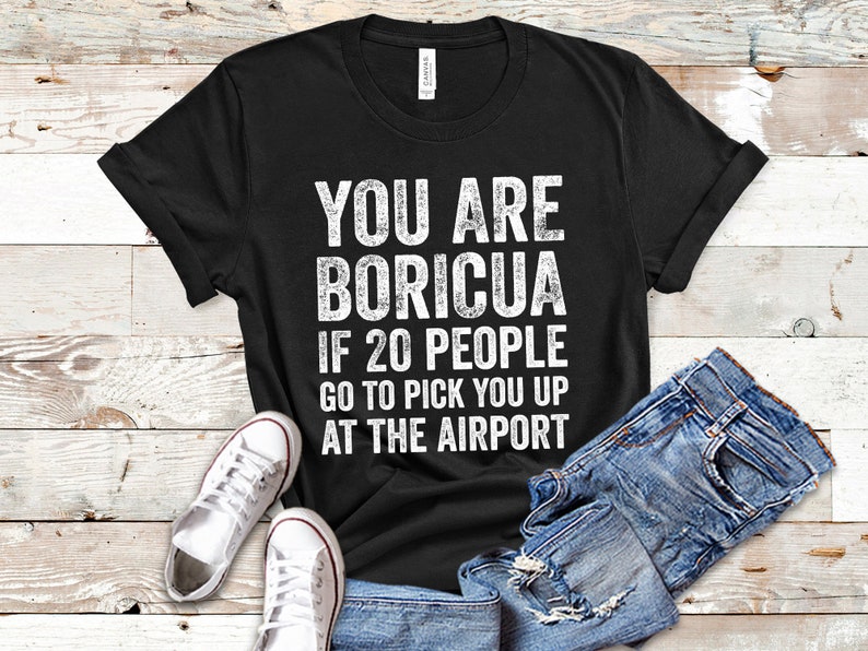 Puede incluir: Camiseta negra con texto blanco: "YOU ARE BORICUA IF 20 PEOPLE GO TO PICK YOU UP AT THE AIRPORT." La camiseta se muestra con vaqueros azules y zapatillas blancas.