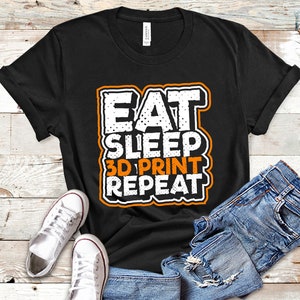 May include: Black t-shirt with the text "EAT SLEEP 3D PRINT REPEAT" in white and orange lettering. The shirt is paired with blue jeans and white sneakers with red accents, all arranged on a light wood-grain surface.