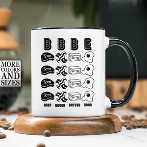 May include: White ceramic mug with a black rim and handle. The mug has a black and white illustration of beef, bacon, butter, and eggs with the text "BBBE" and "BEEF BACON BUTTER EGGS".