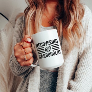 May include: White ceramic mug with black text that reads "RECOVERING CARBOHOLIC" with a graphic of a steak in the middle of the text.