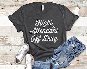 May include: A dark gray t-shirt with the text "Flight Attendant Off Duty" in white with a small airplane graphic. The shirt is laid flat on a woodgrain surface with a pair of white sneakers and blue jeans.