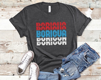 May include: Dark gray t-shirt with the word "BORICUA" in red, white, and blue block letters. The shirt is folded on a light wood surface next to a pair of white sneakers and blue jeans.
