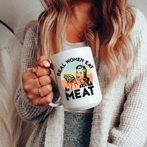 May include: White ceramic mug with a retro-style illustration of a woman holding a piece of meat. The text on the mug reads "Real Women Eat Meat".