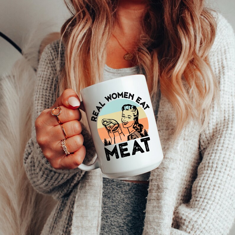 May include: White ceramic mug with a retro-style illustration of a woman holding a piece of meat. The text on the mug reads "Real Women Eat Meat".