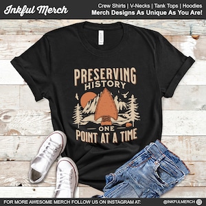 May include: Black t-shirt with a graphic design of a mountain range, a red sun, and an arrowhead. The text "Preserving History One Point At A Time" is printed on the shirt.