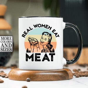 May include: White ceramic mug with a black rim and handle. The mug features a retro-style illustration of a woman holding a piece of meat with the text "Real Women Eat Meat" in a black and white font.