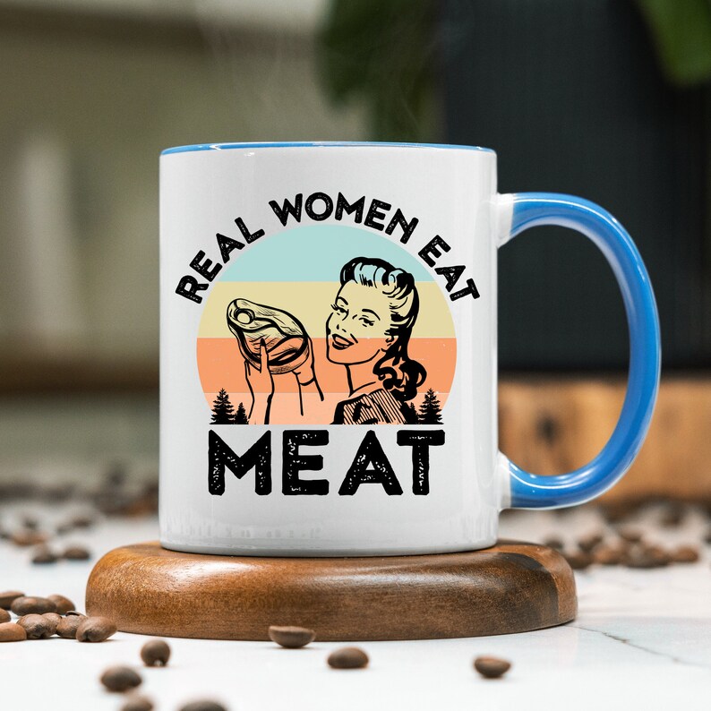 May include: White ceramic mug with a blue rim and handle. The mug features a retro-style illustration of a woman holding a piece of meat with the text "REAL WOMEN EAT MEAT" in a vintage font.