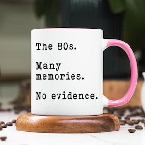 May include: White coffee mug with a pink rim and handle. The mug has black text that reads "The 80s. Many memories. No evidence."